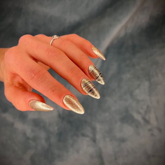 Eine Hand mit golden lackierten Nägeln mit schwarzen Linien. Die Nägel sind spitz und glänzend. Ein Ring ist am Ringfinger.