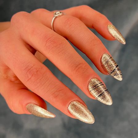 Eine Hand mit goldener, komplizierter Nagelkunst, die vor einem verschwommenen Hintergrund präsentiert wird. Jeder Nagel hat ein einzigartiges Design.