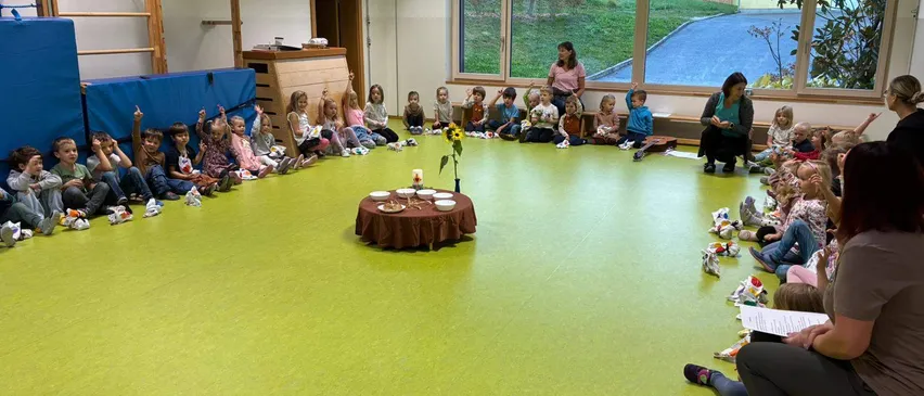 Eine Gruppe von Kindern sitzt im Kreis auf dem Boden um einen kleinen Tisch mit einer Sonnenblume, während zwei Frauen in der Nähe stehen.