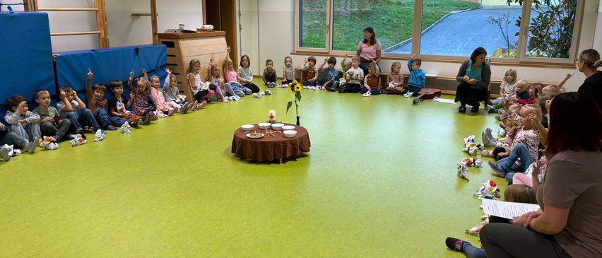 Eine Gruppe von Kindern sitzt im Kreis auf dem Boden um einen kleinen Tisch mit einer Sonnenblume, während zwei Frauen in der Nähe stehen.