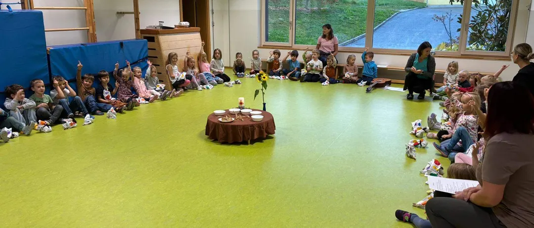 Eine Gruppe von Kindern sitzt im Kreis auf dem Boden um einen kleinen Tisch mit einer Sonnenblume, während zwei Frauen in der Nähe stehen.