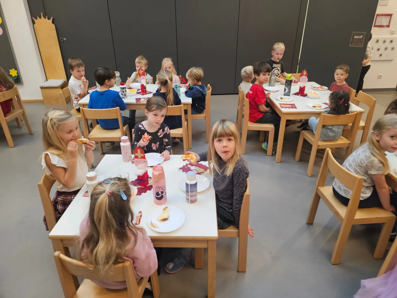 Kinder sitzen in einem Klassenzimmer an Tischen und essen mit Tellern vor sich. Einige haben Getränke auf dem Tisch und andere haben ein rotes Kreuz auf ihren Hemden.