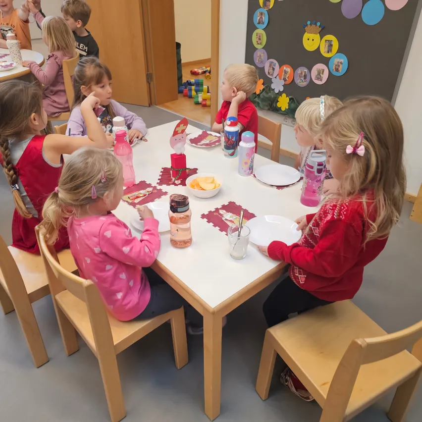 Mehrere junge Kinder sitzen um einen Tisch herum und essen. Sie haben Teller, Gläser und Essen vor sich. Hinter ihnen befindet sich eine Wand mit verschiedenen Dekorationen.