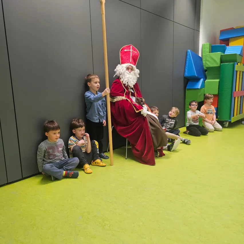 Eine Gruppe von Kindern sitzt um einen Mann, der als Weihnachtsmann verkleidet ist und einen großen Stab hält. Sie sitzen auf einem grünen Boden mit bunten Blöcken im Hintergrund.
