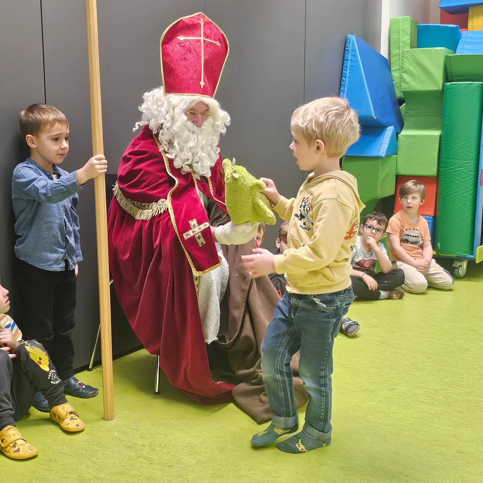 Eine Person in der Kleidung des Heiligen Nikolaus interagiert mit einem jungen Jungen in einem Spielzimmer, umgeben von anderen Kindern.