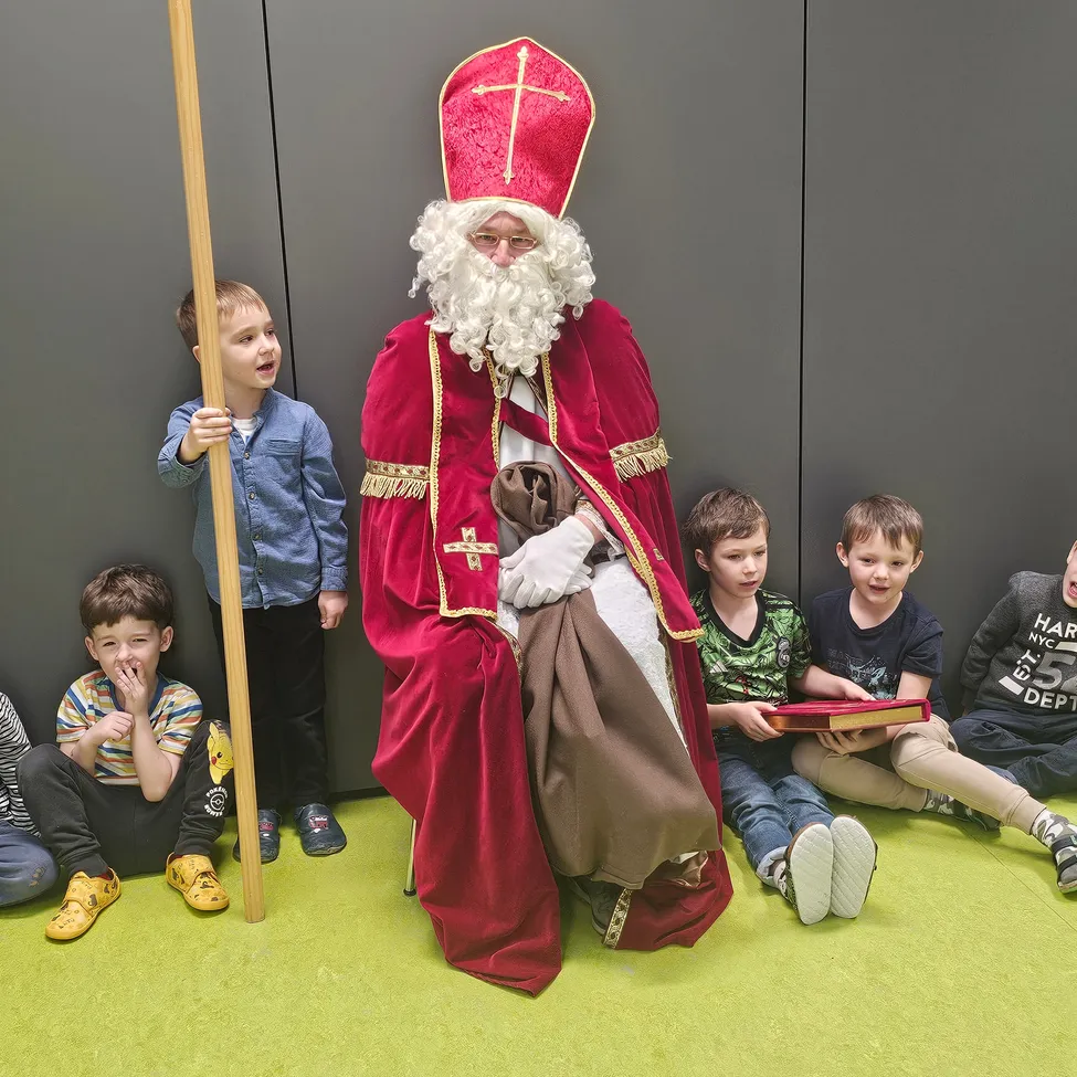 Ein Mann, der als St. Nikolaus verkleidet ist, sitzt umgeben von Kindern. Sie sitzen auf einem grünen Boden, einer hält ein Buch.