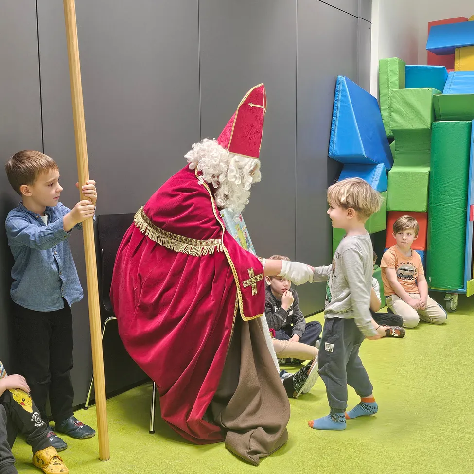 Ein Mann in Bischofskleidung schüttelt einem Jungen die Hand, während ein anderer Junge seinen Stab in einem Raum mit buntem Spielbereich hält.