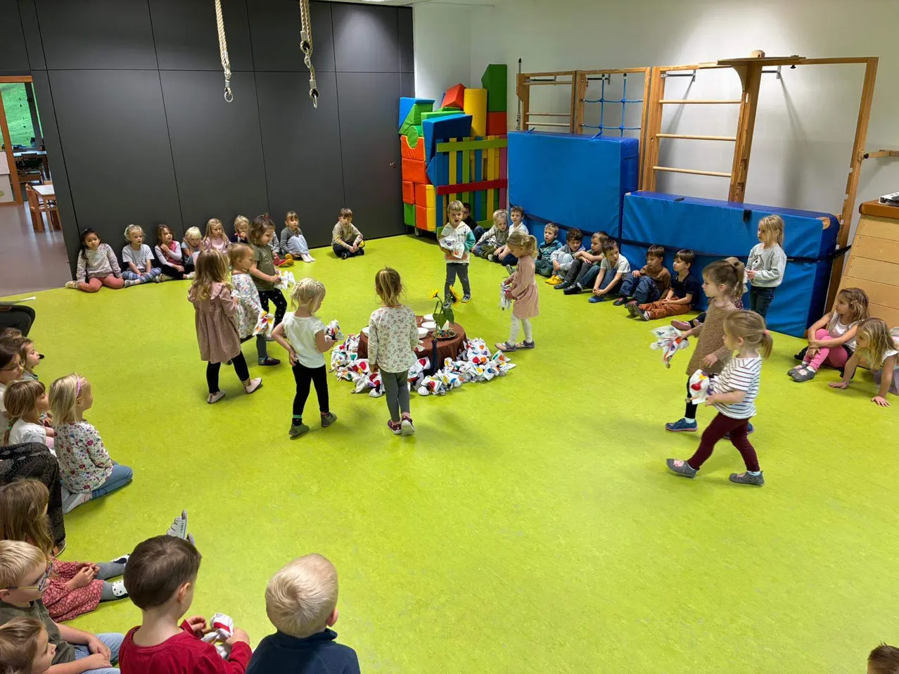 Eine Gruppe von Kindern befindet sich in einem Spielzimmer mit gelbem Boden und nimmt an einer Gruppenaktivität teil. Sie sind von verschiedenen Spielzeugen und bunten Strukturen umgeben.