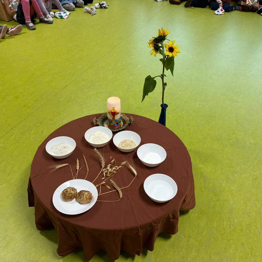 Ein runder Tisch mit einem braunen Tischtuch ist mit Schalen voller Getreide, Brot und einer Kerze besetzt. Sonnenblumen stehen in einer Vase auf dem Tisch. Kinder sitzen auf grünem Boden um den Tisch herum.