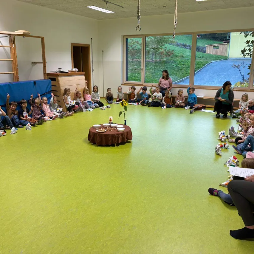 Eine Gruppe von Kindern sitzt im Kreis auf grünem Boden, angeführt von zwei Frauen. Ein kleiner Tisch mit einer Sonnenblume steht in der Mitte, und der Raum hat große Fenster mit Blick auf die Grünflächen draußen.