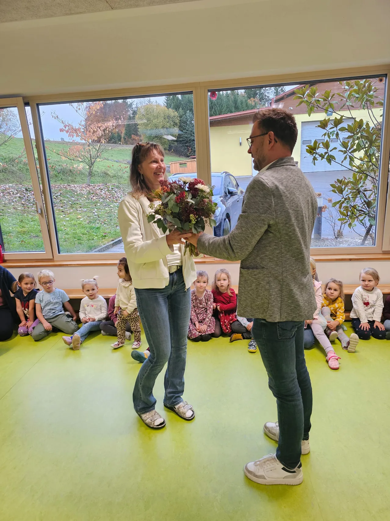 Ein Mann überreicht einer Frau einen Blumenstrauß in einem Raum mit grünem Boden, umgeben von Kindern, die auf dem Boden sitzen. Vor dem Fenster ist ein Auto geparkt.