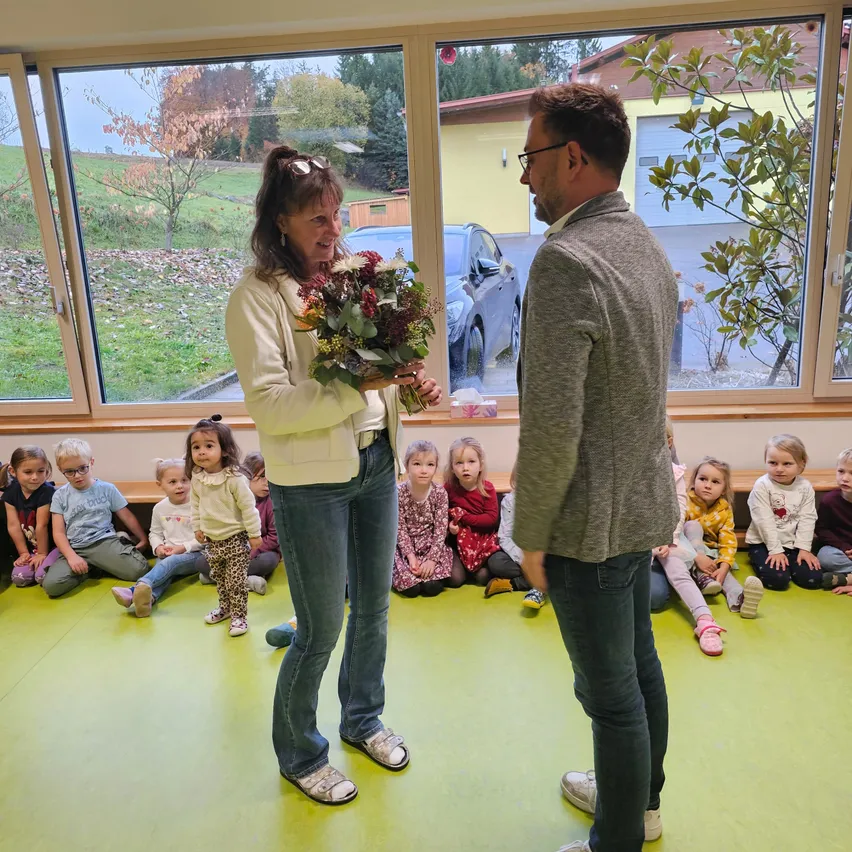 Eine Frau mit Blumen steht neben einem Mann, der eine Gruppe von Kindern auf dem Boden sitzend ansieht, in einem Raum mit großen Fenstern.