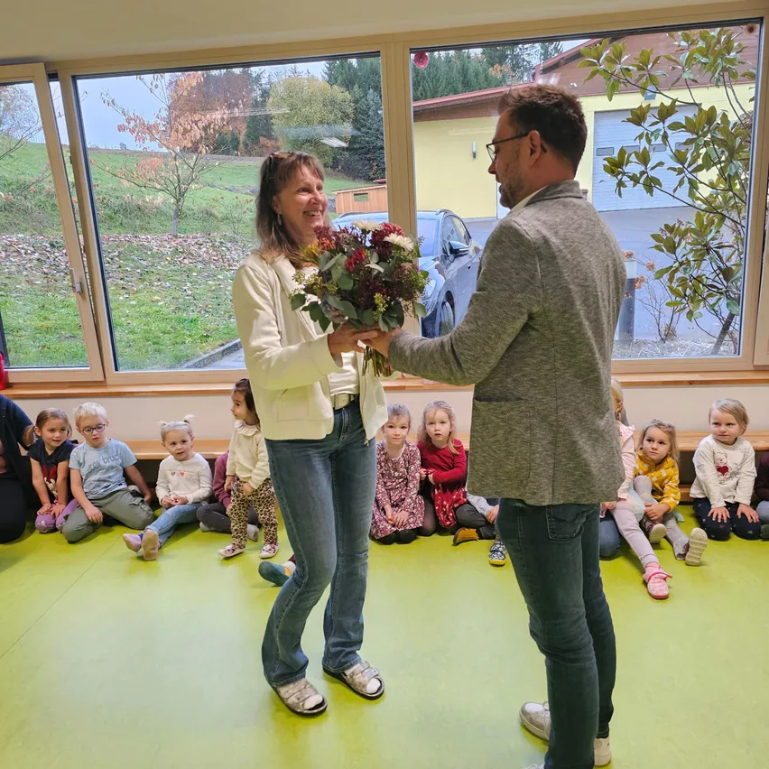 Ein Mann überreicht einer Frau Blumen, während eine Gruppe von Kindern hinter ihnen in einem Klassenzimmer sitzt.