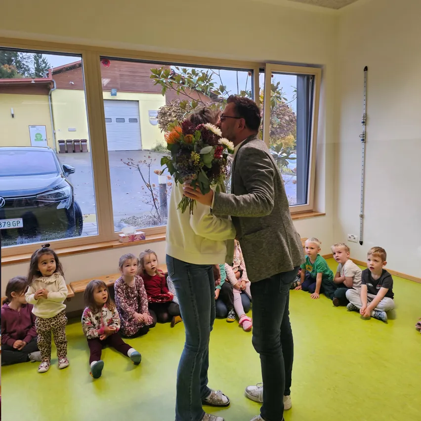 Ein Mann und eine Frau halten Blumen und küssen sich in einem Raum. Kinder sitzen auf dem Boden und beobachten sie. Der Raum hat große Fenster, und ein Auto ist draußen geparkt.