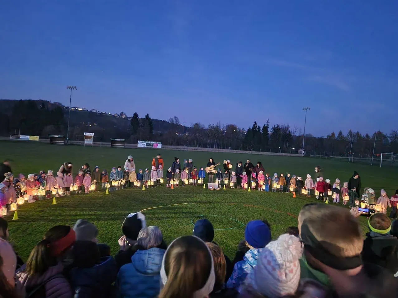 Viele Menschen sind auf einem Rasenfeld bei Dämmerung versammelt, mit Kindern, die brennende Kerzen in einem Kreis halten. Sie sind von Erwachsenen umgeben, einige tragen Mützen und Mäntel. Straßenlaternen und Bäume sind im Hintergrund zu sehen.