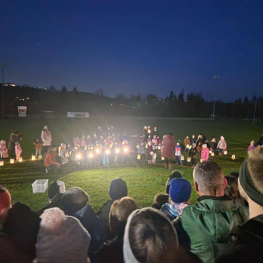 A nighttime gathering on a grassy field, people holding lit candles. Trees and streetlights in the background.