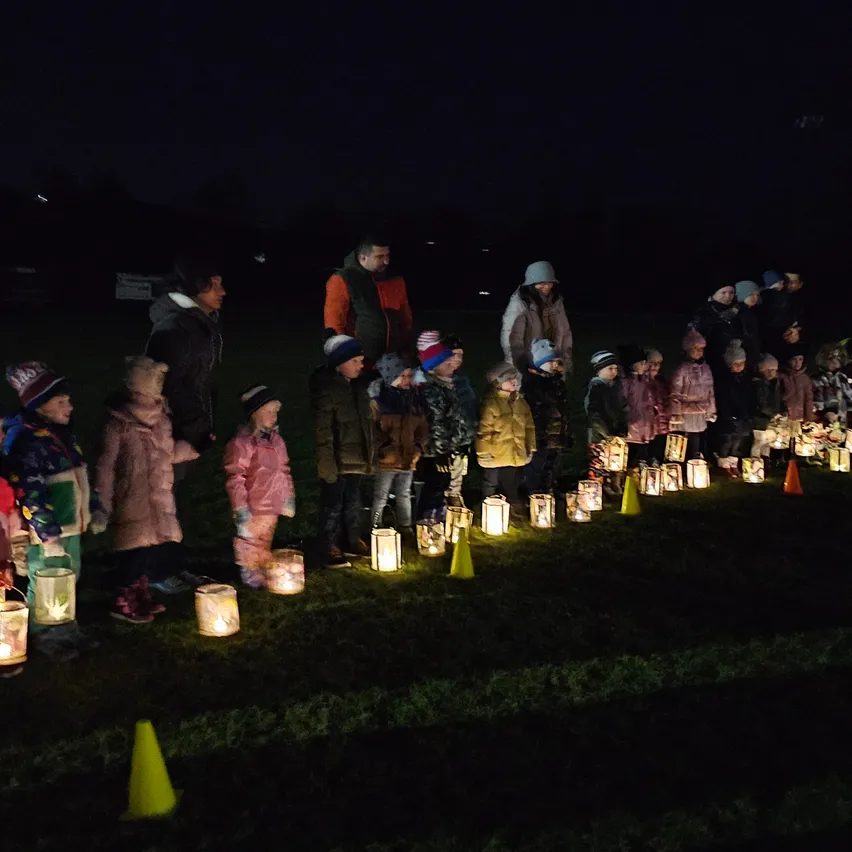 Kinder und Erwachsene stehen in einer Reihe und halten brennende Laternen in der Nacht. Einige tragen Mützen, und der Boden ist mit Kegeln markiert. Bäume und ein Schild sind im Hintergrund zu sehen.