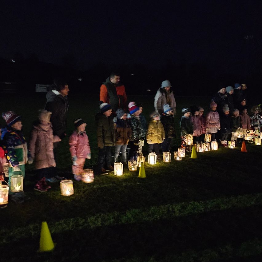 Children and adults stand in a row, holding lit lanterns at night. Some wear hats, and the ground is marked with cones. Trees and a sign are visible in the background.