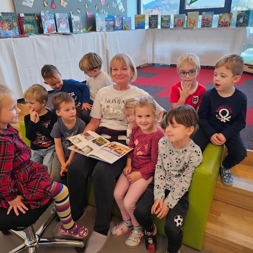 Eine ältere Frau liest ein Buch vor einer Gruppe von Kindern, die um sie herum auf einem grünen Stuhl sitzen, mit Büchern, die auf einem Tisch im Hintergrund ausgestellt sind.