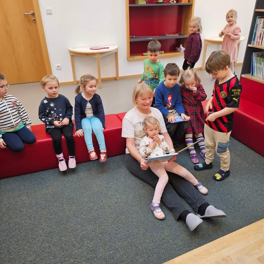 Eine Frau sitzt auf dem Boden und hält ein Baby, während mehrere Kinder auf einer roten Bank um sie herum sitzen und aufmerksam zuhören.