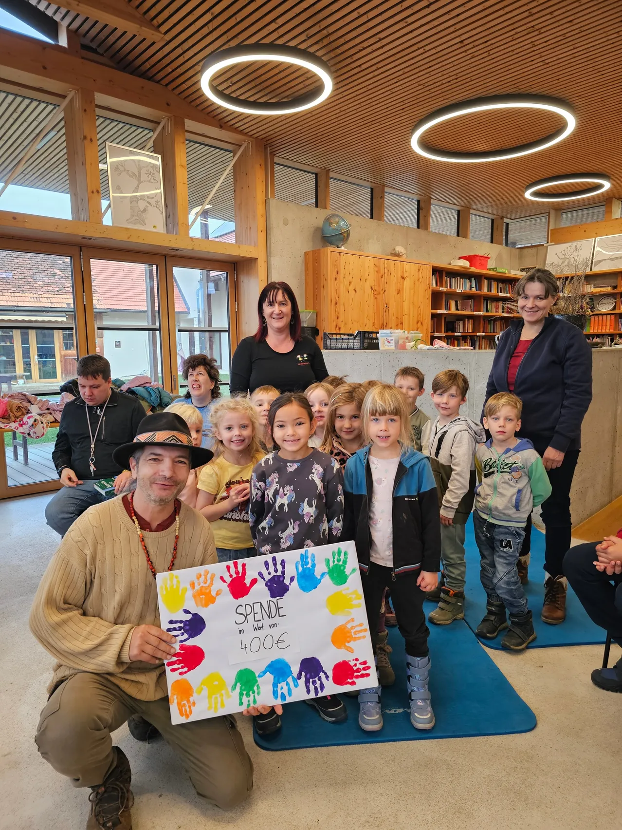 Eine Gruppe von Kindern und Erwachsenen posiert für ein Foto im Klassenzimmer, ein Mann hält ein Schild mit bunten Handabdrücken und dem Text 'Spende in Wert von 4000€'.