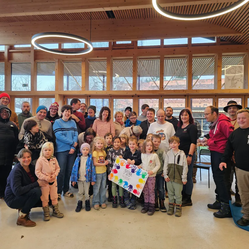 Eine Gruppe von Erwachsenen und Kindern versammelt sich in einem hell erleuchteten Raum mit Holzwänden und großen Fenstern. Sie lächeln und posieren für ein Foto, wobei eine Frau ein farbenfrohes Schild hält.