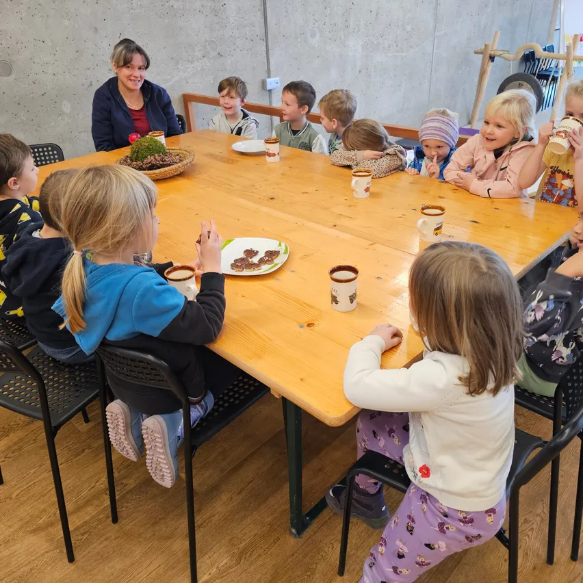 Kinder und ein Erwachsener sitzen um einen Holztisch herum, trinken aus Tassen und essen. Eine Frau steht hinter dem Tisch und lächelt.