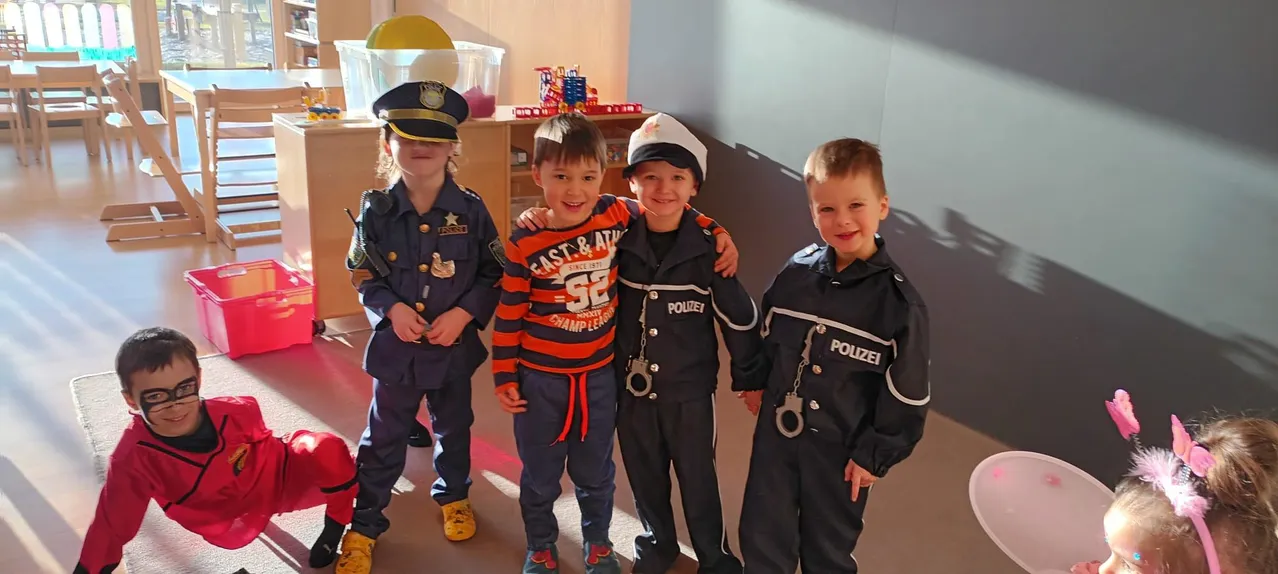 Vier Kinder in Polizeiuniformen posieren für ein Foto. Ein Kind trägt ein gestreiftes Shirt, und ein anderes hat Handschellen. Sie sind in einem Raum mit einem Holzschrank und einer Spielzeugkugel.