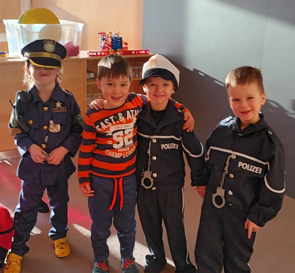 Vier Kinder in Polizeiuniformen posieren für ein Foto. Ein Kind trägt ein gestreiftes Shirt, und ein anderes hat Handschellen. Sie sind in einem Raum mit einem Holzschrank und einer Spielzeugkugel.