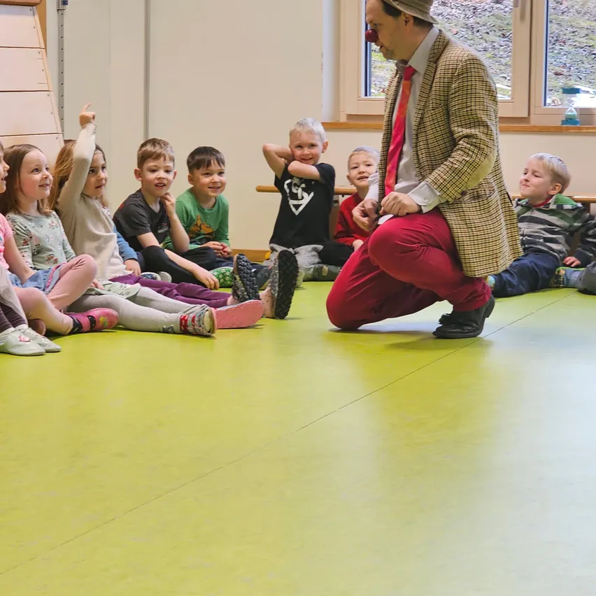 Ein Clown unterhält eine Gruppe von Kindern, die auf dem Boden sitzen. Die Kinder lächeln und schauen den Clown an. Der Raum hat gelben Boden.
