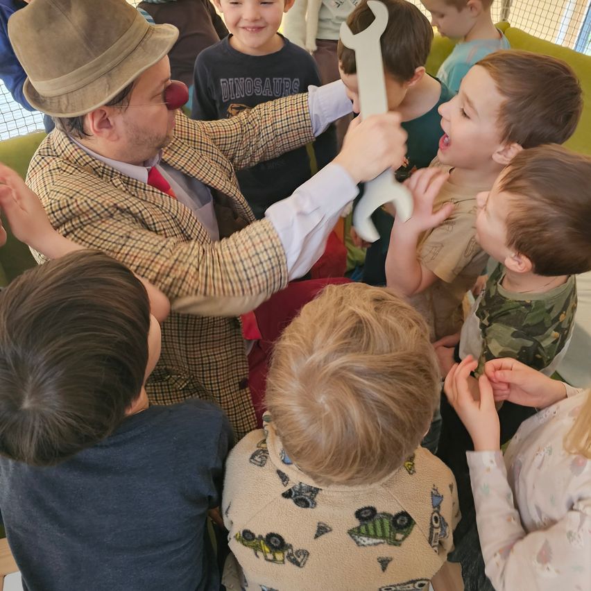 Ein Clown mit einer großen weißen Schraube unterhält eine Gruppe von Kindern, einige greifen danach.
