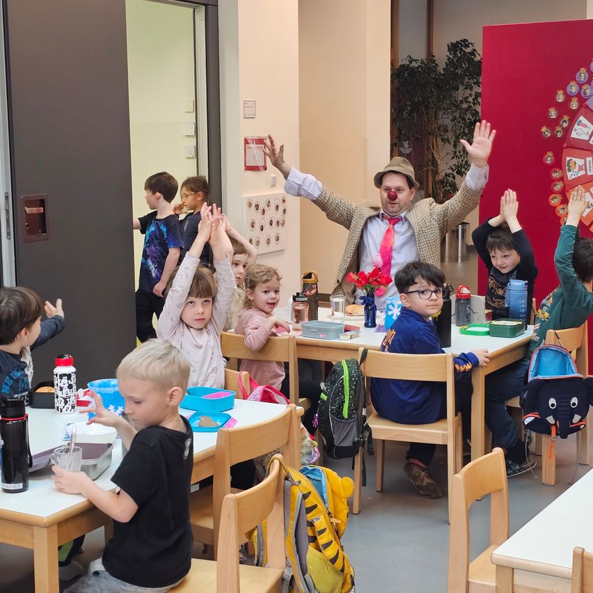 Ein Raum mit Tischen und Stühlen; Kinder sitzen um die Tische herum. Ein Clown steht in der Mitte des Raumes mit erhobenen Händen. Ein Junge in der Nähe der Tür schaut zum Clown.
