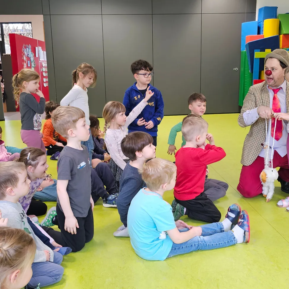 Ein Clown unterhalt eine Gruppe von Kindern in einem Spielzimmer mit bunten Spielzeugen im Hintergrund. Die Kinder sitzen auf dem Boden und horchen aufmerksam zu.