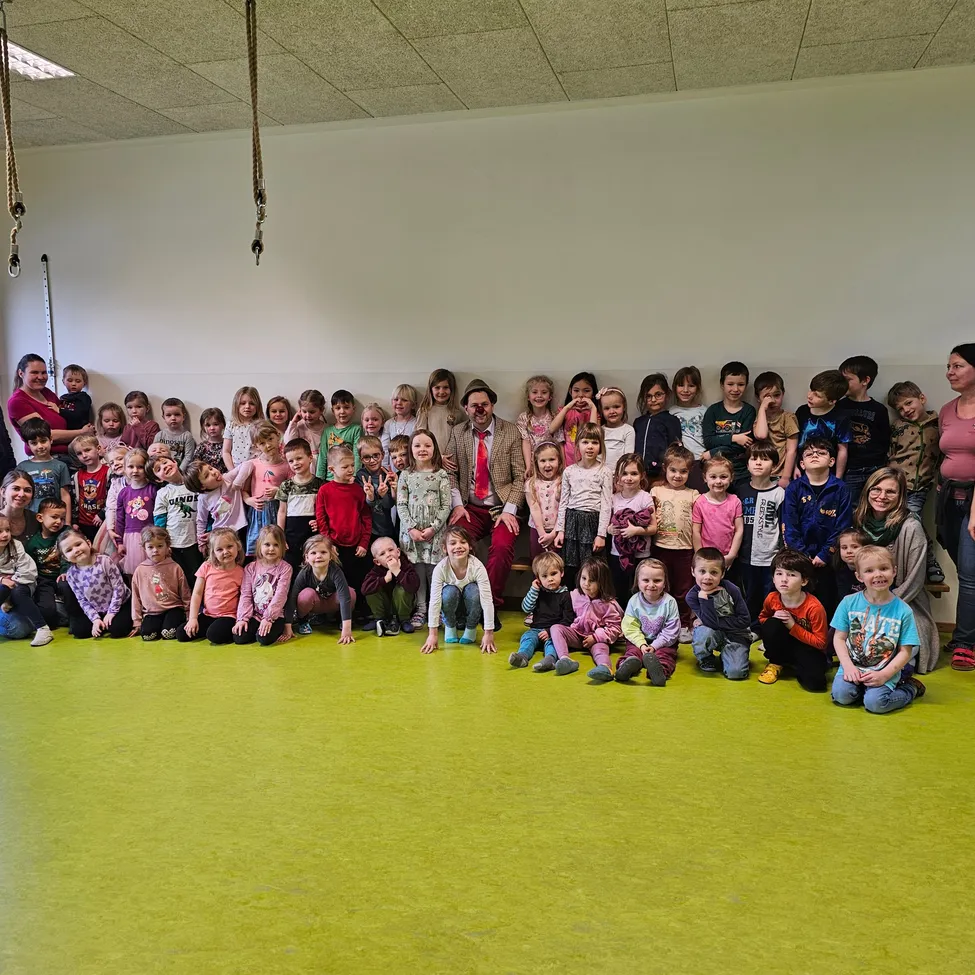 Eine Gruppe von Kindern und Erwachsenen posiert für ein Foto in einem Raum mit grünem Boden und weißer Wand. Die Kinder sitzen und stehen in Reihen, und die Erwachsenen stehen hinter ihnen.