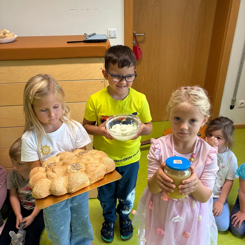 Drei Kinder stehen in einem Raum. Ein Mädchen auf der linken Seite hält einen Tablett mit Brot, ein Junge in der Mitte hält eine Schüssel mit Essen und ein Mädchen auf der rechten Seite hält ein Glas. Hinter ihnen sitzen einige Kinder auf dem Boden.