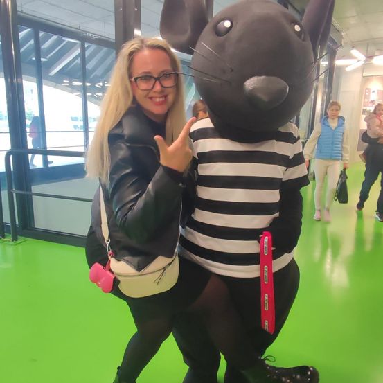 Eine Frau in einer Lederjacke posiert mit einem Maus-Maskottchen in einem Raum mit grünem Boden und Glaswänden.