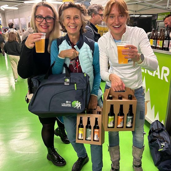 Drei Frauen stehen bei einer Veranstaltung, halten Gläser mit Bier und eine Karton mit Flaschen. Eine von ihnen trägt eine Brille und hält eine große Sporttasche. Der Boden ist grün.