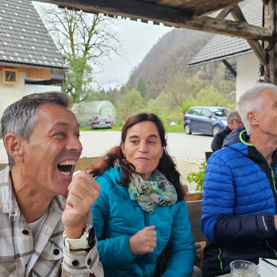 Eine Gruppe von Menschen sitzt unter einem Pavillon, ein Mann lacht, während die anderen ihm zuzuhören scheinen.