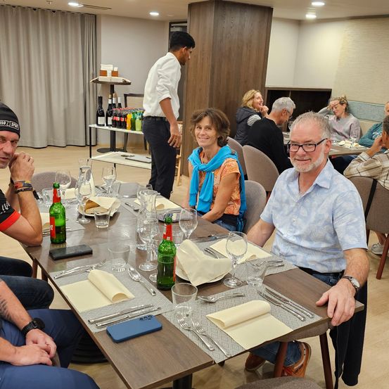 Eine Gruppe von Menschen sitzt an einem Tisch, zwei Männer und eine Frau. Der Tisch ist mit Tellern, Weingläsern und einem Handy gedeckt. Der Mann links trägt einen schwarzen Hut und ein Armband.