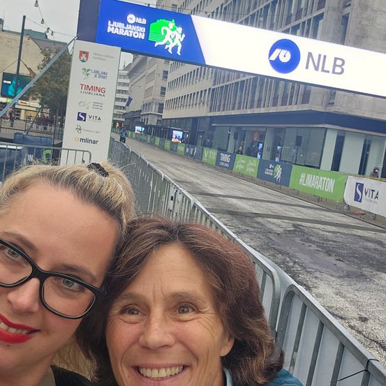 Zwei Frauen stehen vor einer Barriere und lächeln für ein Foto. Im Hintergrund sind ein Marathonbanner und eine Stadtlandschaft zu sehen.