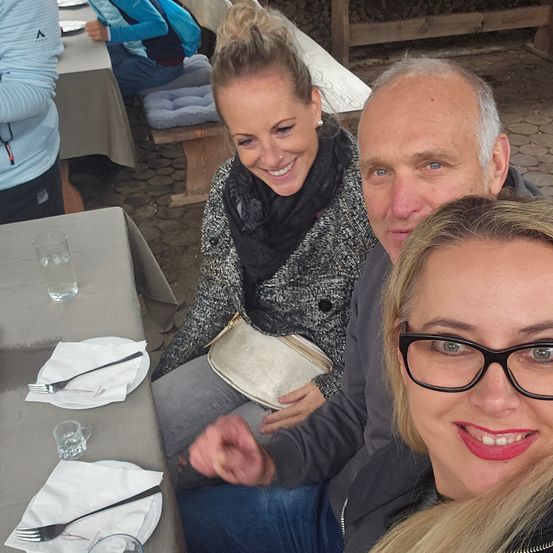 Eine Frau mit einem schwarzen Schal lächelt und posiert für ein Foto mit zwei Männern, die eine Brille tragen.