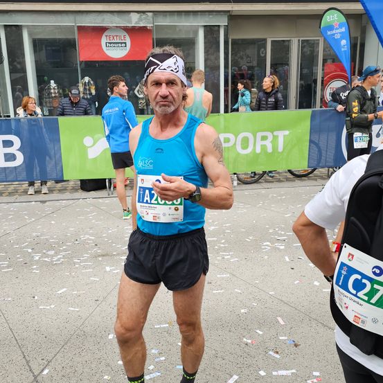 Ein Mann in einem blauen Tanktop und schwarzen Shorts steht auf einer Stadtstraße und hält ein Stück Papier mit seiner Startnummer in der Hand. Der Boden ist mit Konfetti bedeckt. Hinter ihm geht ein anderer Läufer.