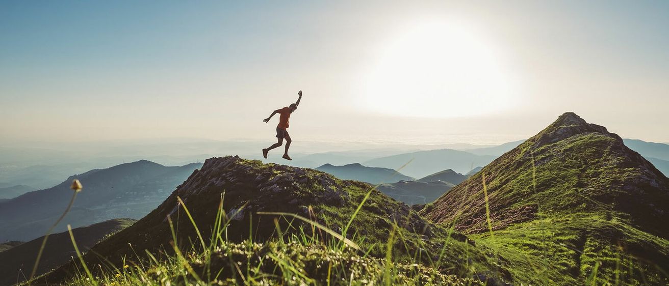 Eine Person in einem roten Shirt springt auf einem felsigen Berggipfel unter einem blauen Himmel und einer untergehenden Sonne.