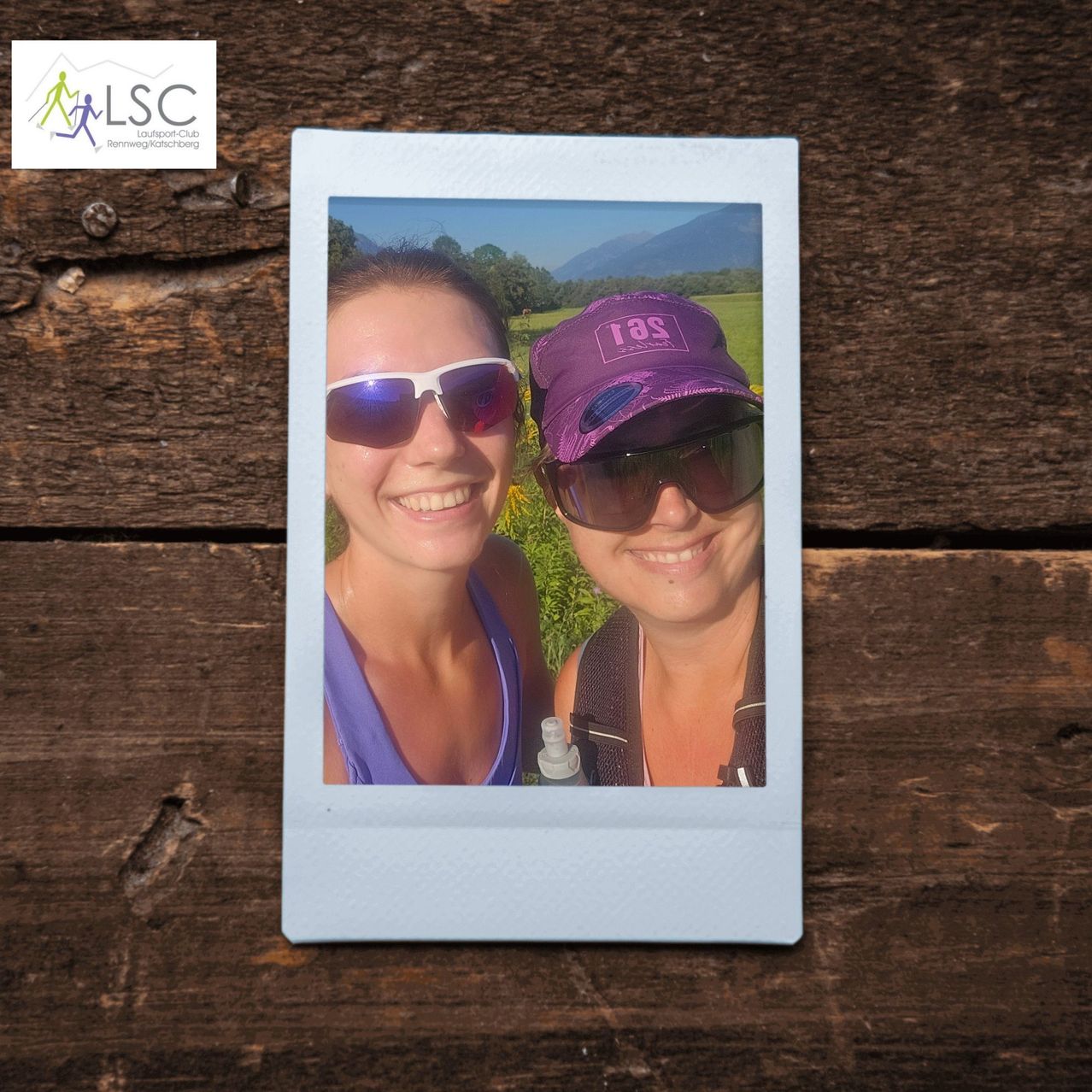 Zwei Frauen mit Sonnenbrillen und Mützen lächeln für ein Foto in einem Gebirgsgebiet. Das Foto ist in einem weißen Quadrat mit einem hölzernen Hintergrund eingerahmt.