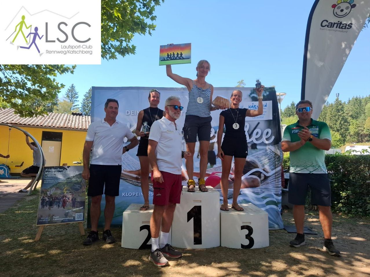 Fünf Personen stehen auf einem Podest mit den Nummern 1, 2 und 3. Die Frau in der Mitte hält eine Regenbogenflagge und eine Medaille. Hinter ihnen ist ein Banner mit dem Text 'LSC' und einem Bild von Läufern.
