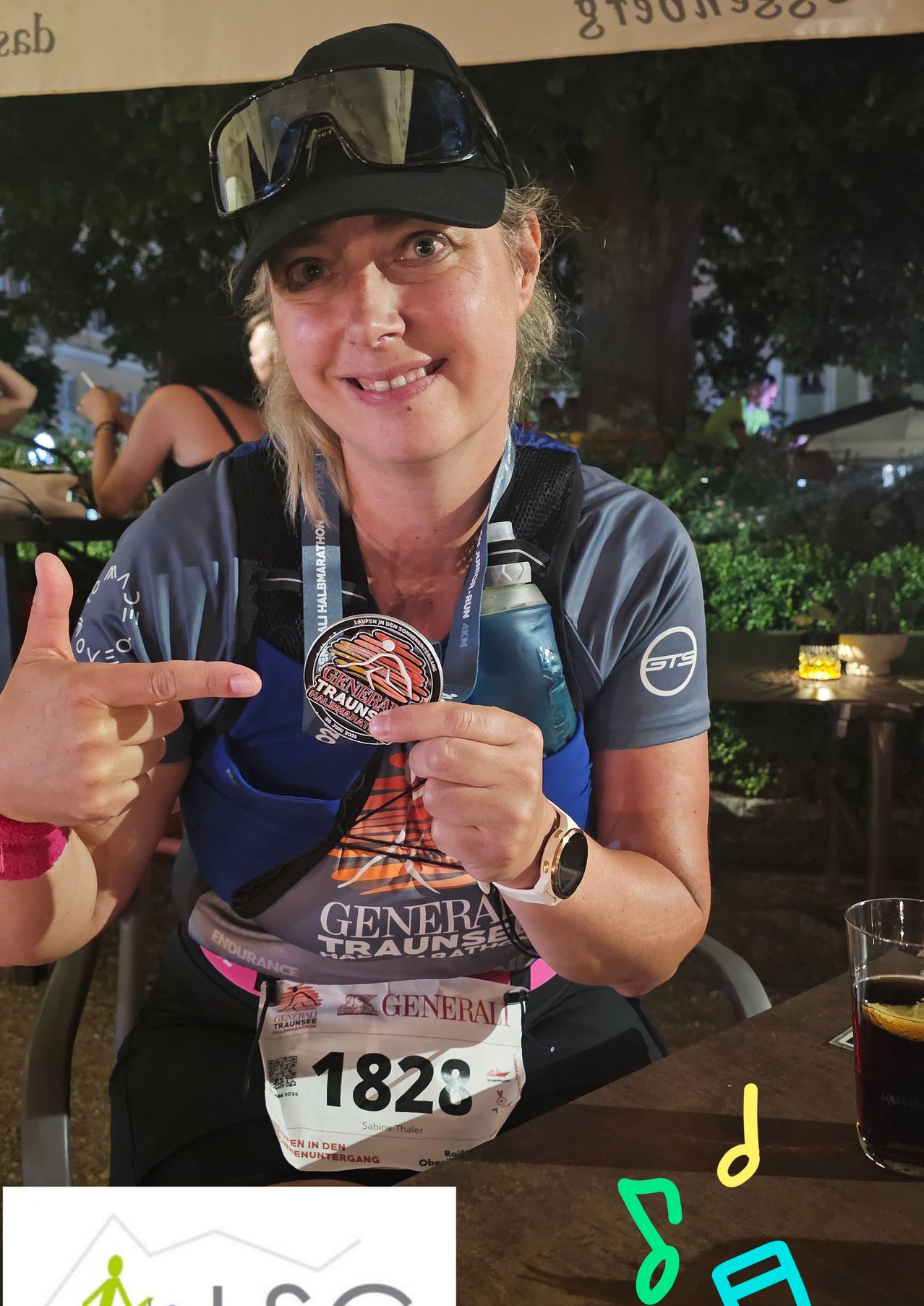 Eine Frau hält eine Medaille und eine Startnummer. Sie gibt einen Daumen hoch. Sie trägt ein Laufshirt und hat eine Wasserflasche. Die Medaille sagt General Traunsee.