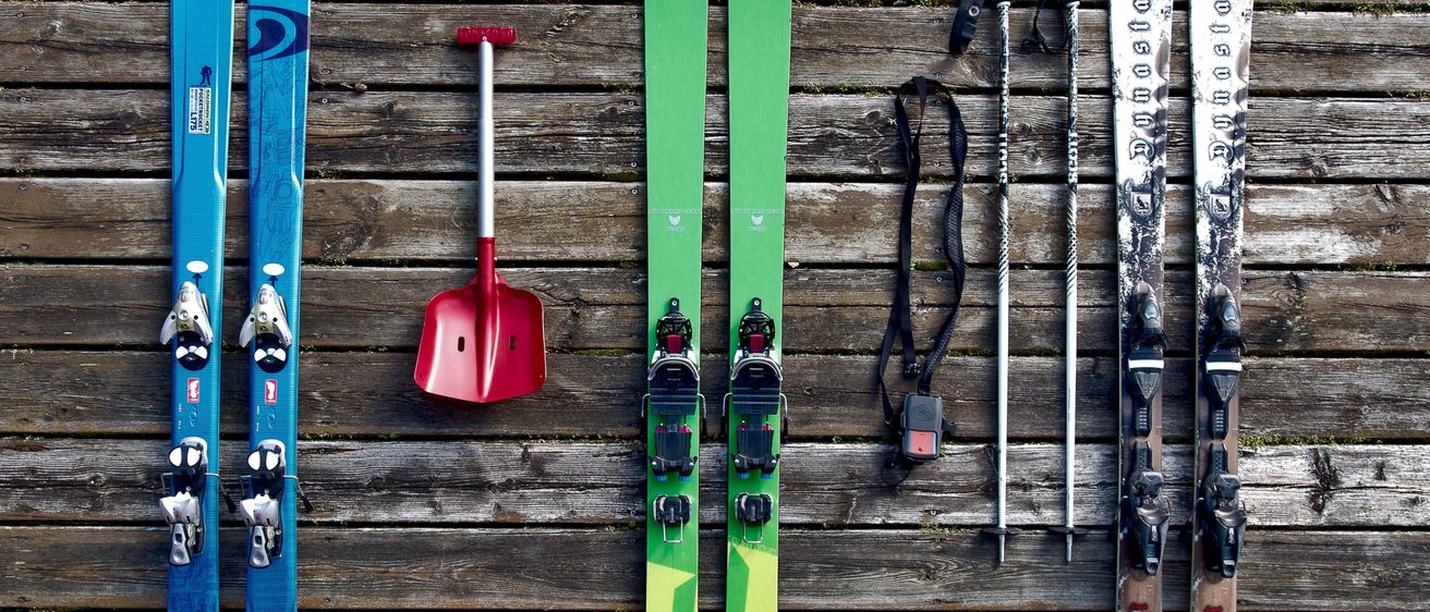 Verschiedene Skier, Skistöcke, eine Schaufel und Skischuhe sind auf einer Holzoberfläche ausgelegt. Die Skier sind in verschiedenen Farben und Größen. Die Skistöcke sind schwarz und silber. Die Schaufel ist rot.
