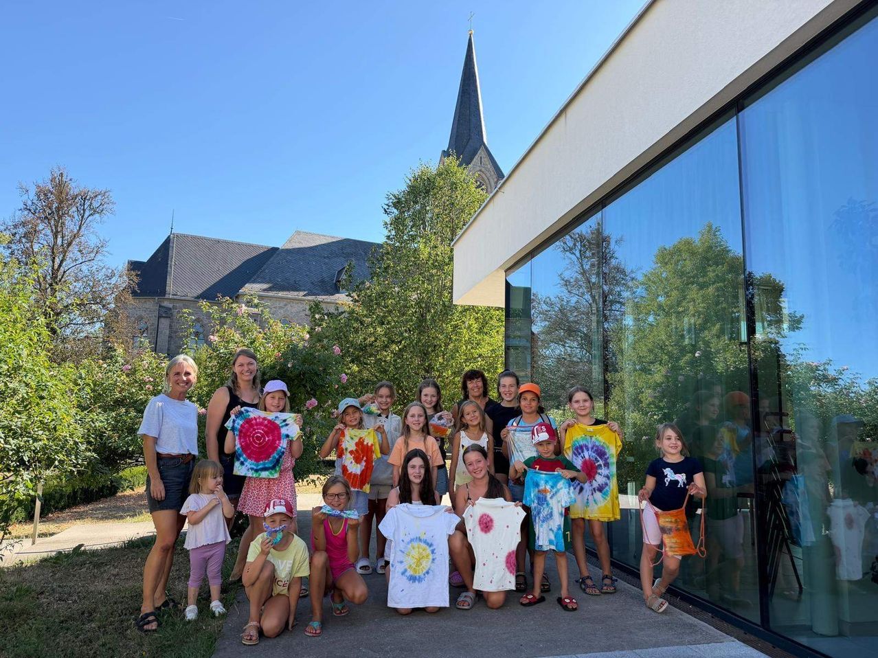 A group of children and adults are posing for a photo in front of a modern building with glass walls, holding tie-dye shirts. Trees and a church with a steeple are in the background.