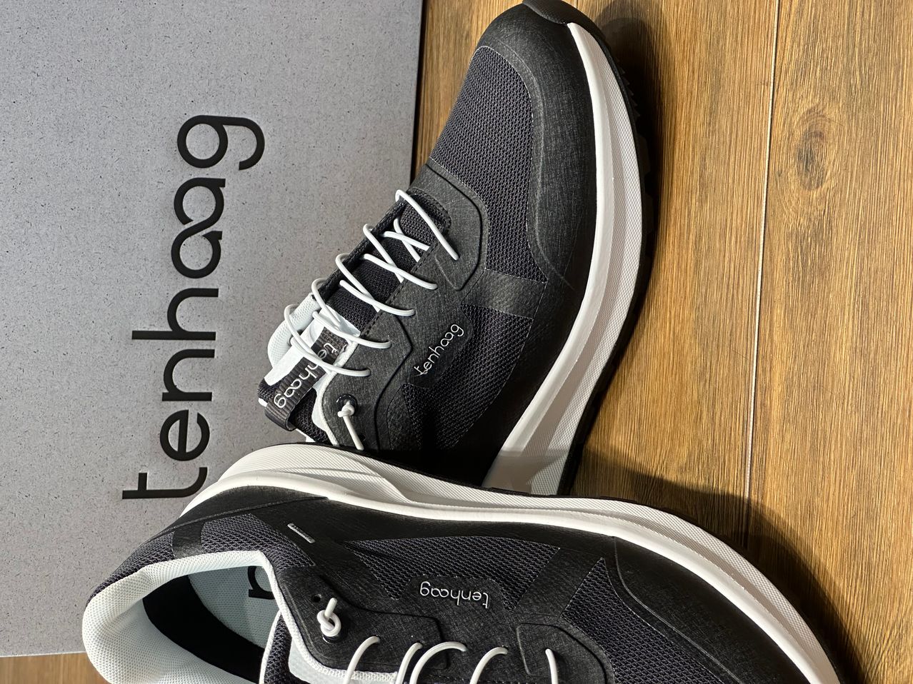 Ein Paar schwarze und weiße Sneaker mit weißen Schnürsenkeln und dem Wort 'tenhaag' auf der Seite werden auf einer Holzoberfläche präsentiert.
