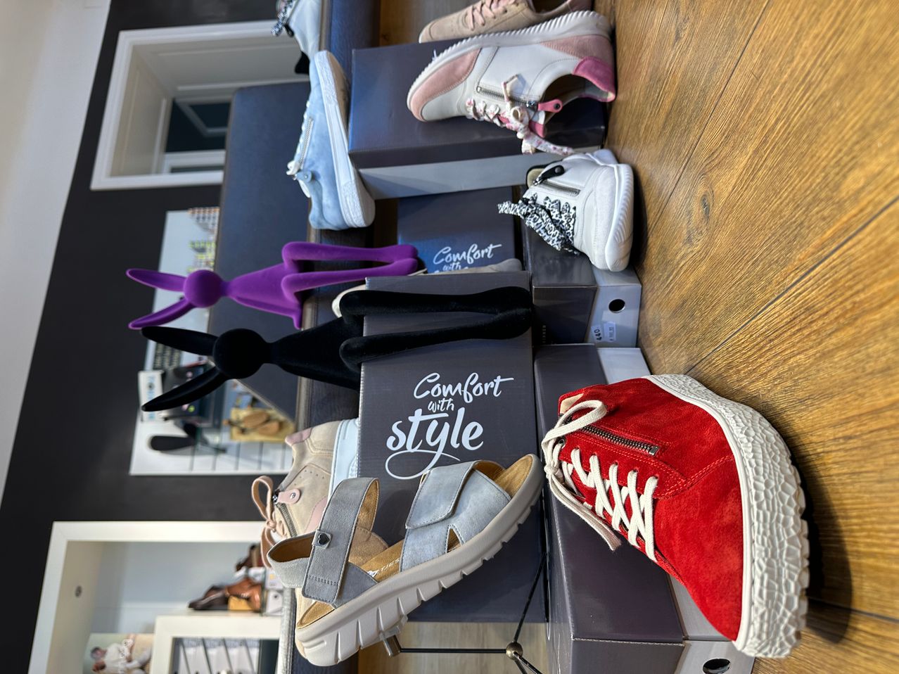 A wooden floor displays various pairs of sneakers, including gray, white, pink, and red, arranged on boxes. A purple figure stands nearby.